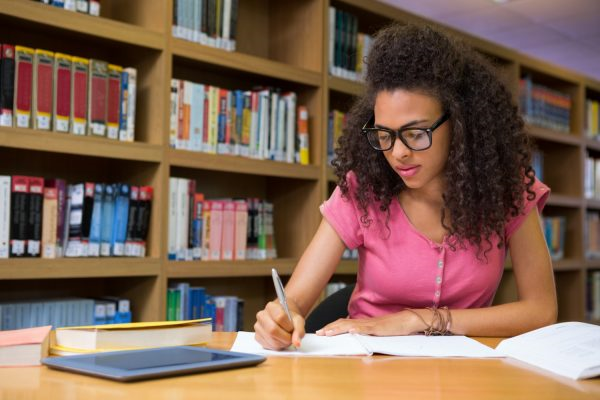 Jovem estudante em uma biblioteca fazendo anotações em um caderno, enquanto estuda quais matérias caem no ENEM. à sua frente, alguns livros e um tablet sobre a mesa.