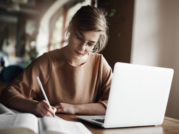 Mulher em uma mesa de estudos, utilizando um notebook e um livro para pesquisar possíveis temas para redação do ENEM. Ela também utiliza uma caneta para registrar as informações em uma folha de papel.