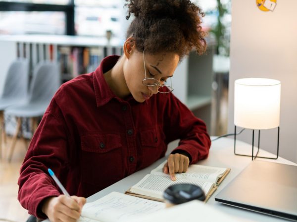 Mulher jovem estudante sentada em sua mesa, utilizando um dos métodos de estudos para absorver melhor a matéria. Ela está lendo um livro enquanto faz anotações em um caderno.